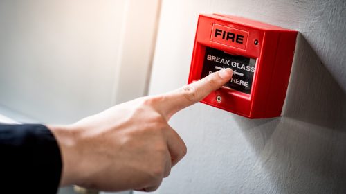 Male hand pointing at red fire alarm switch on concrete wall in office building. Industrial fire warning system equipment for emergency.