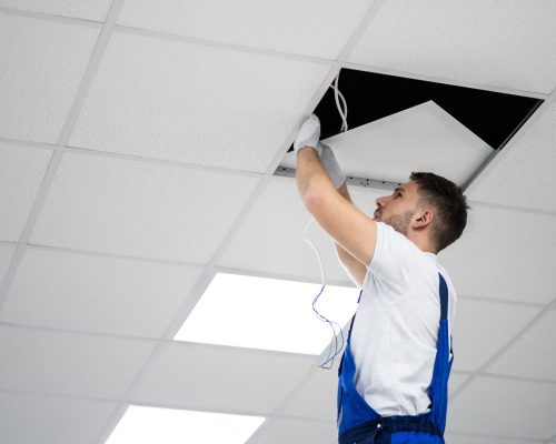 Full Length Portrait Of Electrician On Stepladder Installs Lighting To The Ceiling In Office