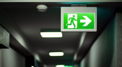 Green fire exit sign on hallway aside of smoke alarm device. Fir