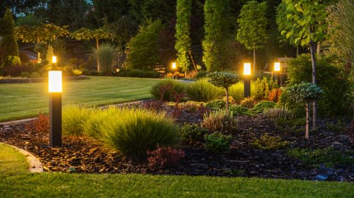 Panoramic Photo of LED Light Posts Illuminated Backyard Garden During Night Hours. Modern Backyard Outdoor Lighting Systems.